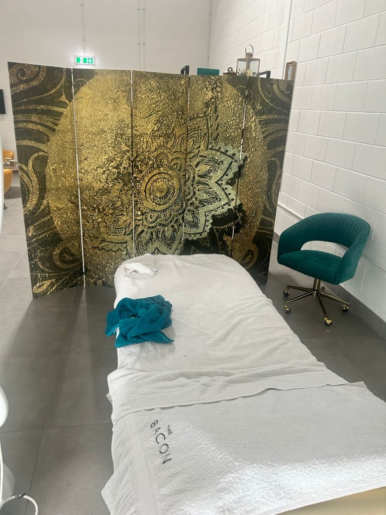 Treatment bed with white towel and teal towel in front of a gold patterned room divider and teal chair in clinic room
