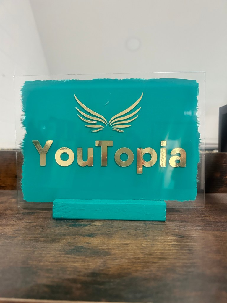 Teal YouTopia sign with gold logo and wings on a wooden shelf