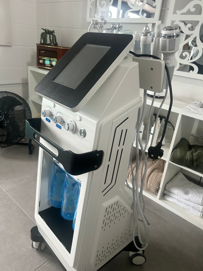 Close-up of a white cosmetic machine with blue liquid containers and various attachments in a clinic treatment room